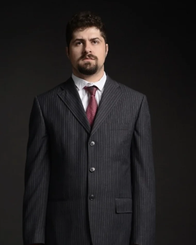 Nikola Dragašević u prugastom odelu, sa koferom u ruci, na tamnoj pozadini.